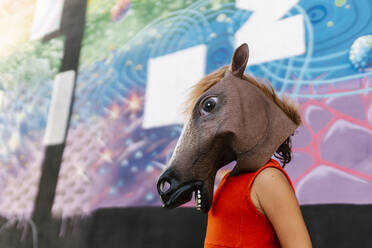 Little girl with a horse's head and a red dress next to a graffiti wall - EGAF00647