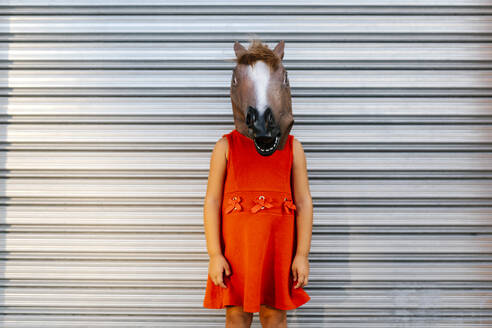 Kleines Mädchen mit einem Pferdekopf und einem roten Kleid vor einem Metalltor - EGAF00650