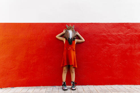 Kleines Mädchen mit einem Pferdekopf und einem roten Kleid Lening auf einer rot-weißen Wand - EGAF00653