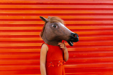 Little girl with a horse's head and a red dress standing in front of red roller shutter - EGAF00655