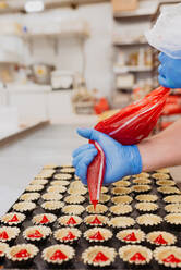 Anonymous confectioner in latex gloves squeezing tasty sweet jelly from bag into small pastry cases on blurred background of bakery kitchen - ADSF10555