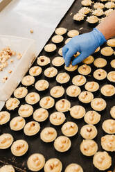 Von oben anonymer Angestellter in Handschuhen, der frische Haselnüsse auf das leckere süße Gebäck auf dem Tablett legt, während er in der Bäckerei arbeitet - ADSF10556