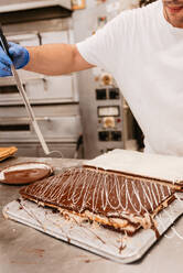 Ein Mann in Latexhandschuhen und Uniform dekoriert eine leckere Torte mit weißen Sahnetupfen bei der Arbeit in einer Bäckerei - ADSF10558