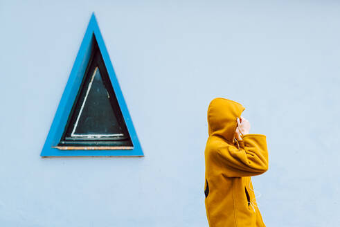 Seitenansicht einer nicht erkennbaren Frau, die die Kapuze einer gelben Jacke zurechtrückt, während sie vor einer grauen Gebäudewand mit dreieckigem Fenster steht - ADSF10579