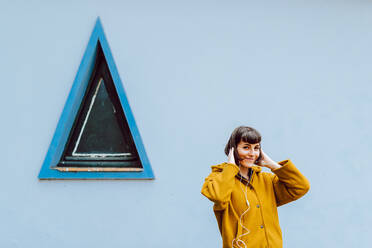 Cheerful female in yellow warm jacket smiling and touching hair while listening to music near gray building with triangle window - ADSF10580