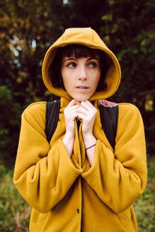 Fröstelnde Frau, die sich in einen gelben Kapuzenmantel hüllt und wegschaut, während sie auf einem unscharfen Naturhintergrund an einem kalten Tag steht - ADSF10581