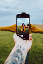 Crop tätowierte Hand mit Smartphone zu fotografieren junge Frau mit ausgestreckten Armen in grünem Feld in der Landschaft - ADSF10583