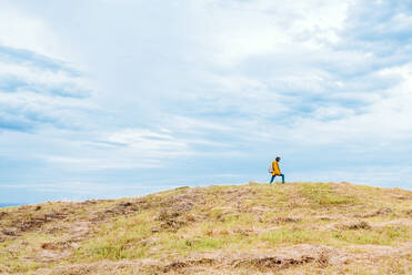 Side view of unrecognizable woman walking on top of grassy hill against cloudy sky in countryside - ADSF10584