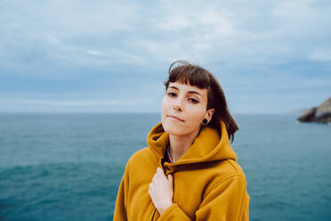 Hübsche Frau in gelber warmer Jacke schaut in die Kamera, während sie gegen das plätschernde Meer und den bedeckten Himmel in der Natur steht - ADSF10585