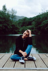 Unique female hipster with short hairstyle in casual clothing sitting with crossed legs on wooden pier of pond with green trees on blurred background - ADSF10591