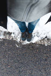 Draufsicht auf eine nicht erkennbare Person in Jeans und Trekkingschuhen, die auf einer asphaltierten, verschneiten Straße bei Tag steht - ADSF10610
