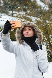 Lächelnde Frau mit Nasenpiercing in weißer Winterjacke mit Kapuze und schwarzen Armstulpen, die ein Selfie mit dem Handy macht, während sie im Winter in der Natur steht, mit Büschen im Hintergrund - ADSF10611
