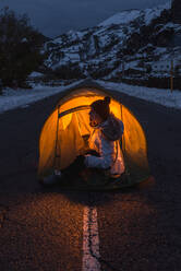 Side view of female in winter clothes sitting at entrance of tourist tent illuminated from inside standing in middle of asphalt road in mountain village at evening - ADSF10613