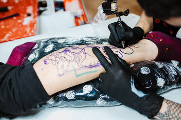 From above anonymous cropped tattooist in latex gloves using tattoo machine to make sketch on leg of crop client in modern salon - ADSF10616