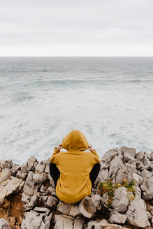 Rückenansicht des Reisenden in gelben warmen Kapuzenpulli sitzen allein auf felsigen Ufer Blick auf schäumende Wellen auf bewölktem Tag - ADSF10626
