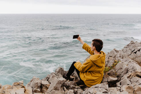 Rückenansicht einer kurzhaarigen, nicht erkennbaren Frau in einem gelben Sweatshirt, die am felsigen Meeresufer über dem schäumenden, wogenden Meer sitzt und ein Selfie mit ihrem Handy an einem grauen, bewölkten Tag macht - ADSF10627