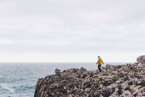 Seitenansicht einer Person im gelben Kapuzenpulli, die an einem grauen, bewölkten Tag auf ein steiniges, von schäumenden Wellen umgebenes Ufer tritt - ADSF10628