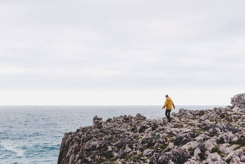 Side view of person in yellow hoodie stepping on stony shore surrounded by foamy waves on gray cloudy day - ADSF10628