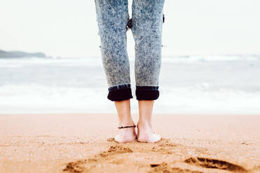 Rückenansicht der Beine einer barfuß lebenden Frau in Jeans, die am Sandstrand mit stürmischem Meer und grauem Himmel im Hintergrund steht - ADSF10638