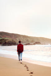 Rückenansicht einer jungen, kurzhaarigen Hipster-Frau in kariertem Hemd und Jeans, die am Strand spazieren geht und Fußabdrücke im nassen Sand hinterlässt - ADSF10642