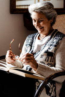 Entspannte reife Frau mit grauem Haar und freundlichem Lächeln, die in einem Sessel am Fenster ein Buch liest und eine Brille in der Hand hält - ADSF10644