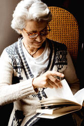 Content woman with gray hair and kind smile reading book by window in light day - ADSF10645