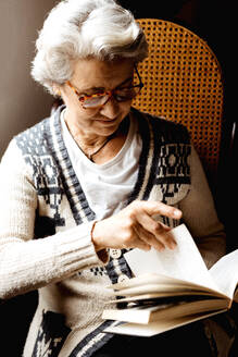 Zufriedene Frau mit grauem Haar und freundlichem Lächeln, die bei hellem Tag am Fenster ein Buch liest - ADSF10645