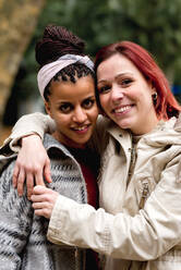 Stilvolles schönes multiethnisches Paar lächelnder Frauen, die sich umarmen und in die Kamera schauen, im Park - ADSF10648