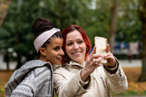 Hübsches multiethnisches Paar lächelnder Frauen, die nah beieinander bleiben und ein Selfie mit dem Handy im Herbstpark machen - ADSF10649