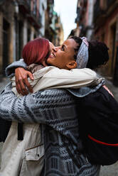 Side view of lovely multiethnic smiling women hugging at meeting in narrow street with old houses - ADSF10657