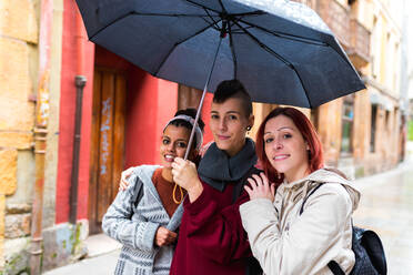 Inhalt trendige multiethnische Frauen, die unter einem großen schwarzen Regenschirm spazieren gehen und in die Kamera schauen, vor dem Hintergrund roter, alter, schäbiger Wände - ADSF10659