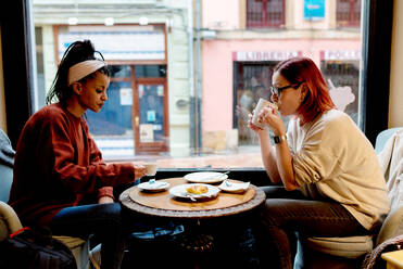 Seitenansicht von stilvollen enthusiastischen multiethnischen Freundinnen, die am Tisch in der Nähe des Fensters in einem Café sitzen und reden und trinken - ADSF10660