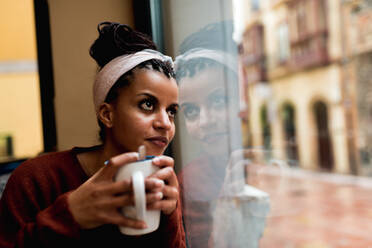 Nachdenkliche, charmante afroamerikanische Frau, die sich an ein Glasfenster lehnt, Kaffee trinkt und verträumt wegschaut - ADSF10661