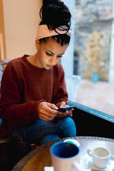 Thoughtful stylish African American woman surfing mobile phone sitting with crossed legs at table with coffee in cafe - ADSF10662