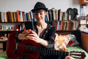 Lächelnde Frau in modischer Kleidung und Hut sitzt auf dem Bett mit Akustikgitarre und liest Nachrichten auf dem Handy, während sie am Wochenende chillt - ADSF10663