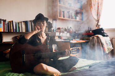 Entspannte Frau in stilvoller Kleidung und Hut sitzt mit gekreuzten Beinen auf dem Bett und raucht mit geschlossenen Augen eine Zigarette - ADSF10664