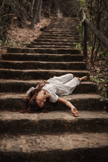 Von oben zarte, charmante junge Frau in weißem Kleid, die verletzlich auf einer Treppe in einem herbstlichen Park liegt und wegschaut - ADSF10669