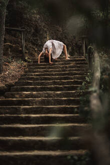 Junge Frau in weißem Kleid steht in seltsamen Brücke Pose stützt sich auf die Hände auf der Treppe tun Yoga-Übung im grünen Park - ADSF10673