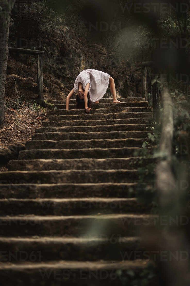 young female in white dress standing in weird bridge pose leaning on hands on stairs doing yoga exercise in green park ADSF10673