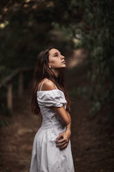 Side view of tender gorgeous young woman in white dress embracing shoulders standing in calm park and looking away - ADSF10675