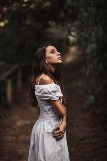 Seitenansicht einer zarten, wunderschönen jungen Frau in weißem Kleid, die ihre Schultern umarmt und in einem ruhigen Park steht und wegschaut - ADSF10675