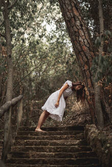 Von unten geheimnisvolle ruhige Frau in weißem Kleid, die sich mit dem Kopf auf einen Baumstamm stützt, der auf einer Treppe in einem ruhigen herbstlichen Park steht - ADSF10678