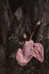 From above of afraid tender young female in pink dress sitting on aged big tree in autumnal forest looking over shoulder - ADSF10685