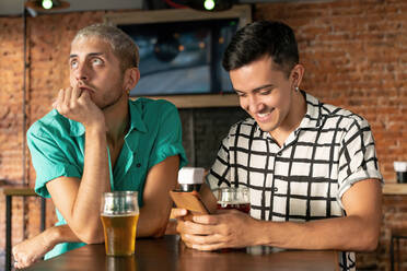 Gelangweilter schwuler Mann, der mit seinem Partner in einer Bar sitzt und ein Mobiltelefon benutzt - SPCF00843