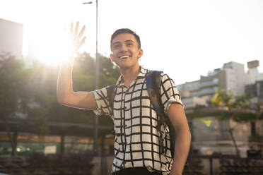 Smiling young gay man waving while standing in city against sky - SPCF00851