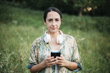 Woman standing with smart phone and headphones in public park - XLGF00453