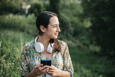 Woman holding smart phone while looking away at public park - XLGF00460