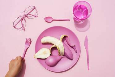 Hand of baby girl picking up fork beside plastic plate with pink-colored pear and banana - GEMF04034