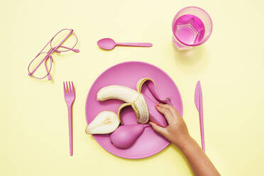 Hand of baby girl picking up pink-colored banana from plastic plate - GEMF04037