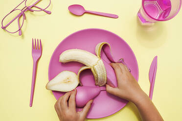 Hand of baby girl picking up pink-colored pear and banana from plastic plate - GEMF04038